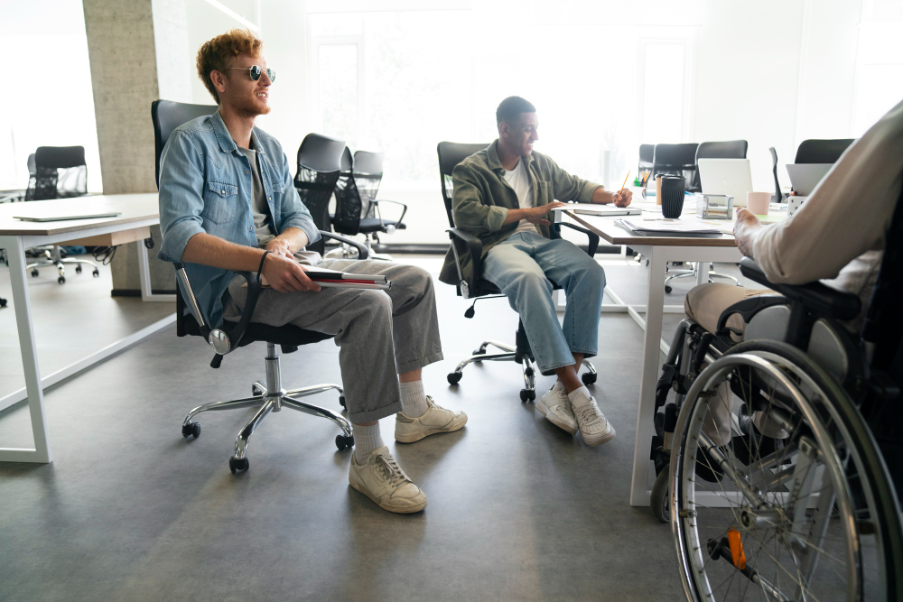 tres personas trabajando en un escritorio. A la izquierda un joven con discapacidad visual utilizando anteojos oscuros, al fondo otro joven trabajando con una notebook y por ultimo un joven en silla de ruedas mirando hacia ellos